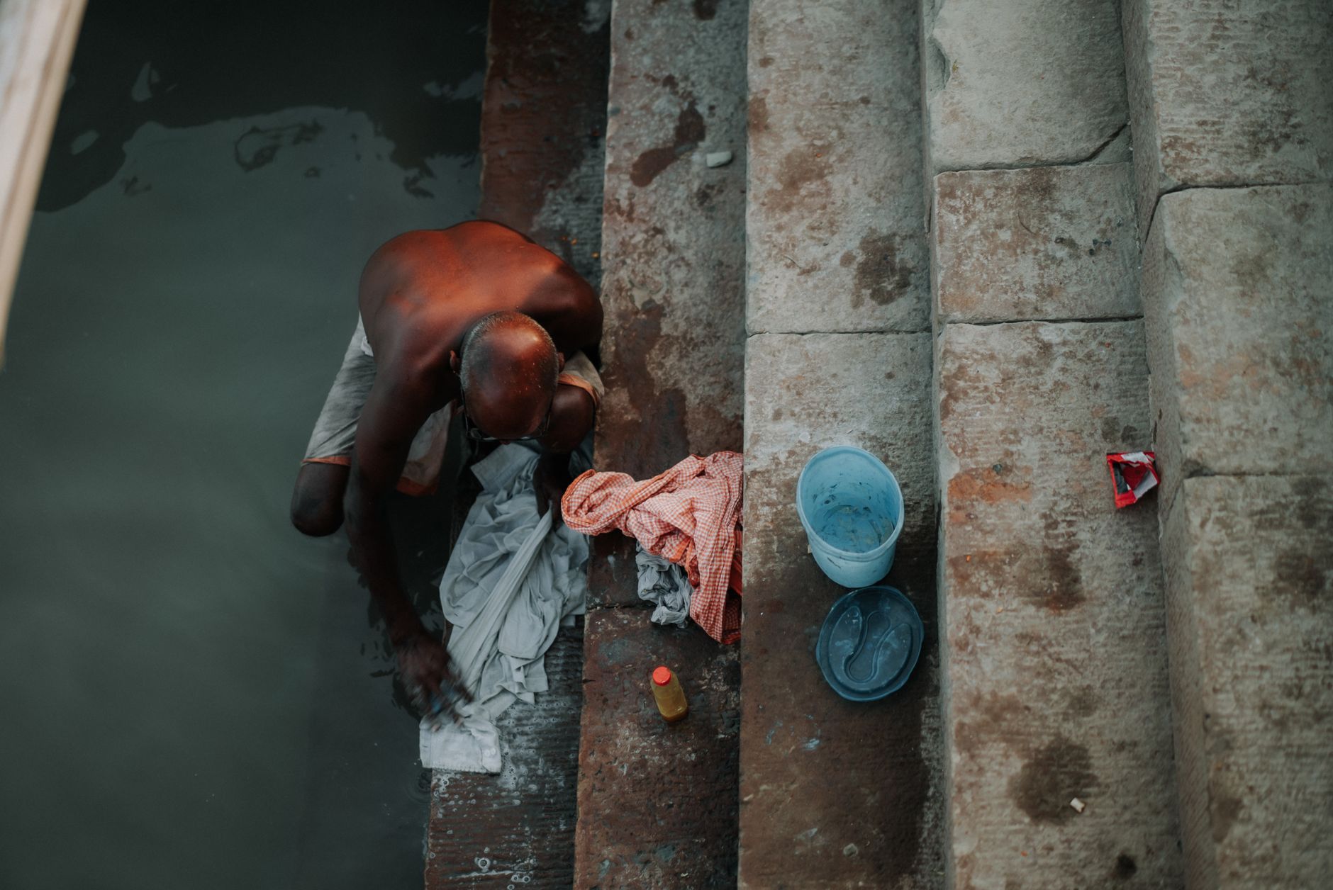 A man washes clothes on river steps with buckets and detergent nearby.