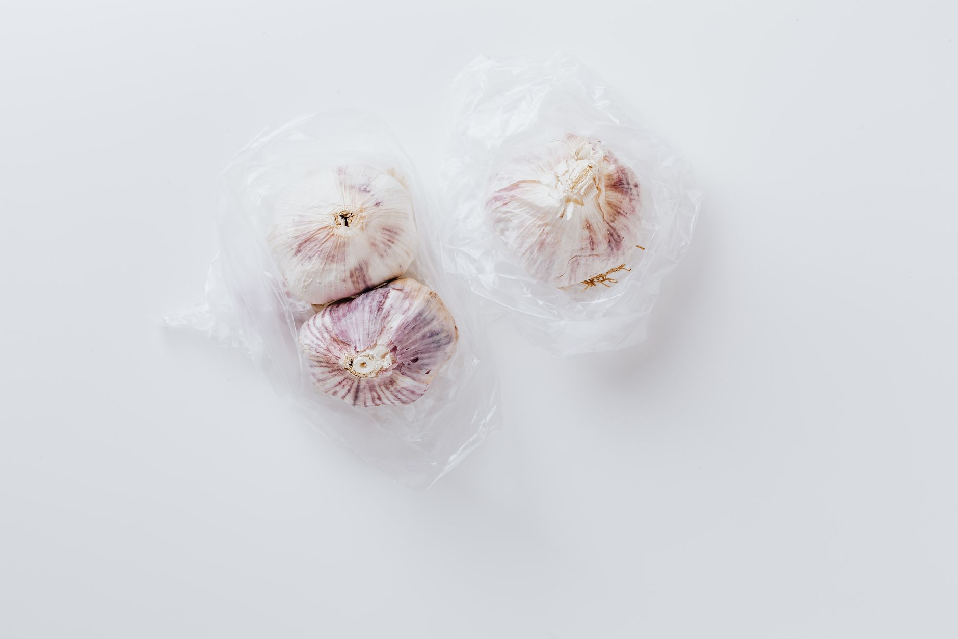 Minimalist image of fresh garlic bulbs wrapped in plastic on a clean white background.